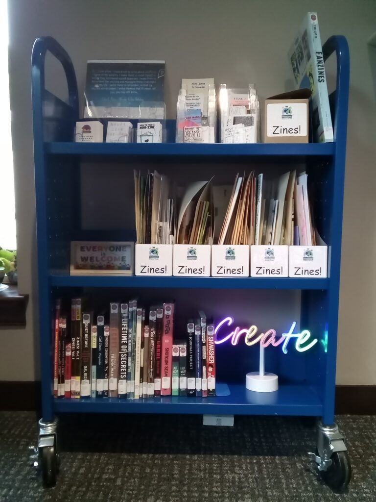 3-shelved library cart on wheels with boxes of zines.