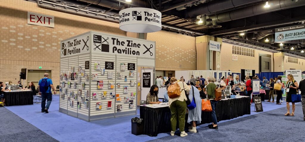 People standing at a table looking at free zines. In the background is a large cube that displays zines and a sign that says Zine Pavilion.